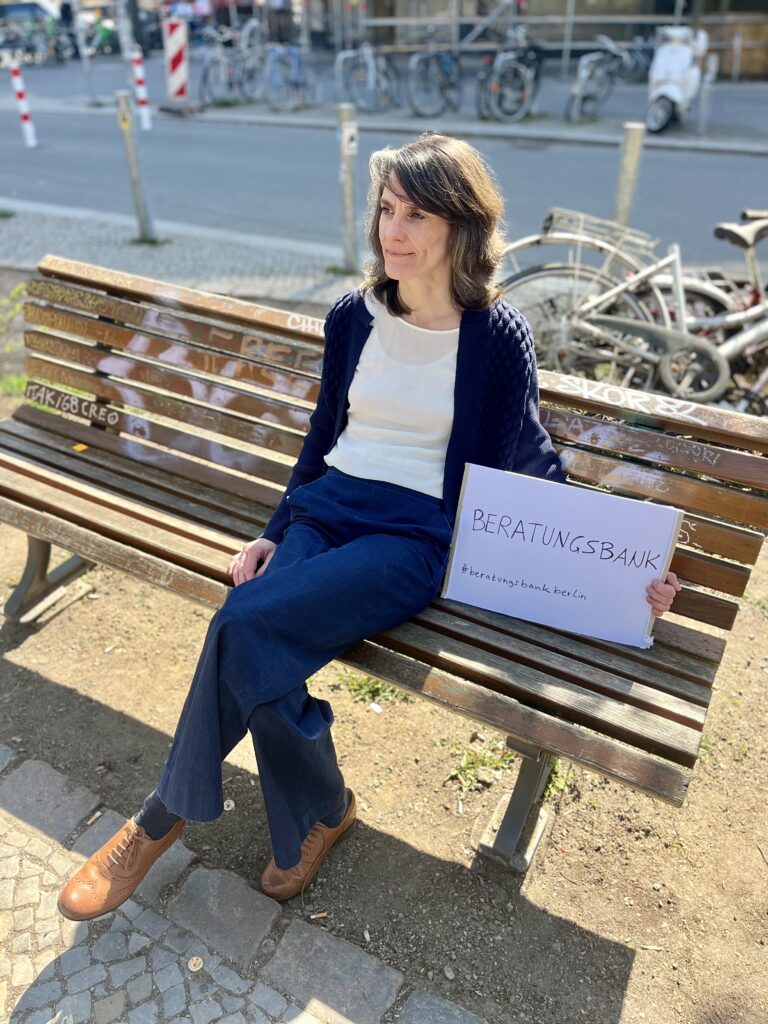 Anna Wolf sitzt auf einer Holzbank im Stadtraum von Neukölln und hält ein Schild mit der Aufschrift „Beratungsbank“.
