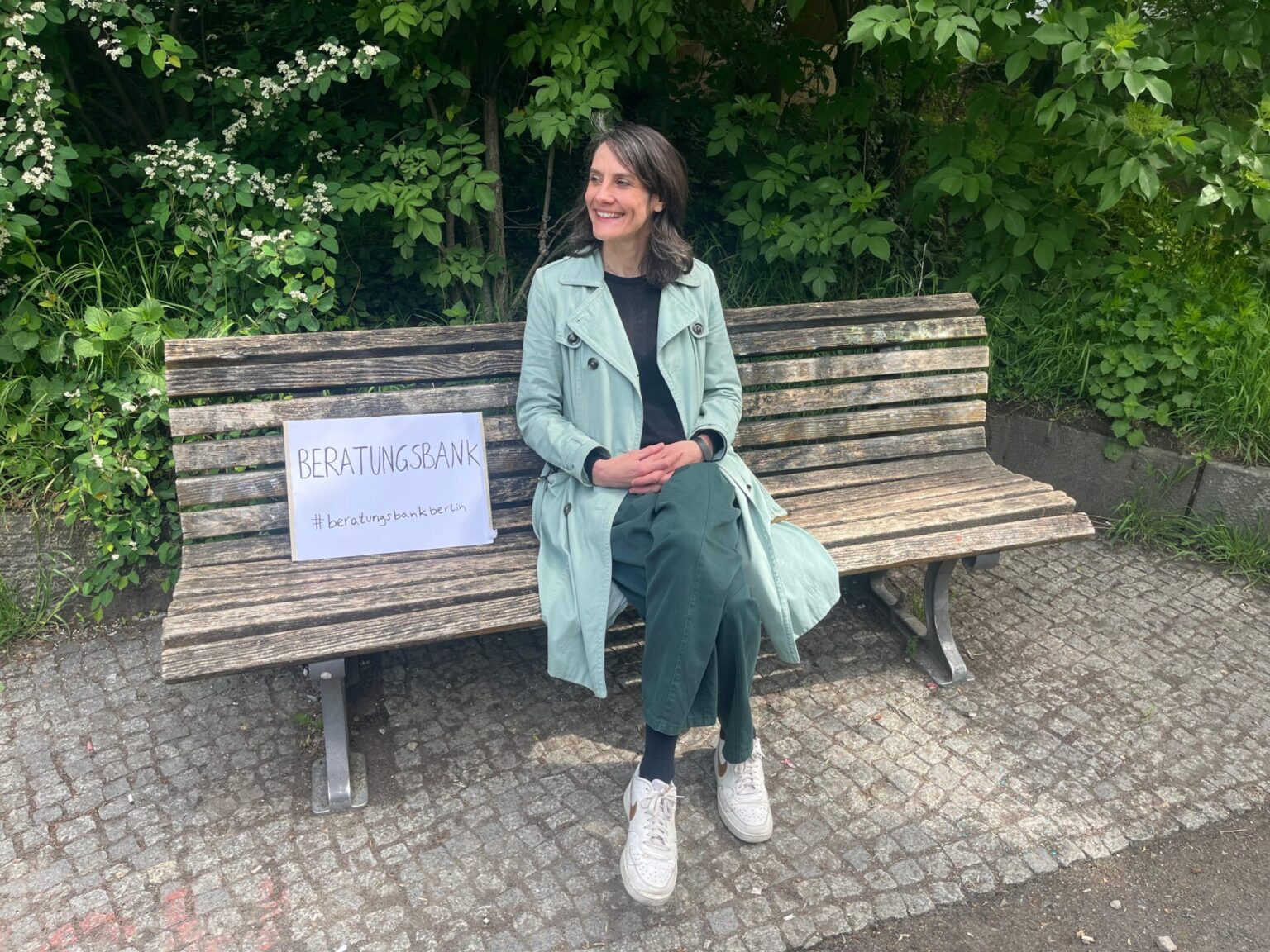Frau in mintgrünem Mantel sitzt lächelnd auf einer Parkbank neben einem Schild mit der Aufschrift „Beratungsbank #beratungsbankberlin", umgeben von grünen Büschen.