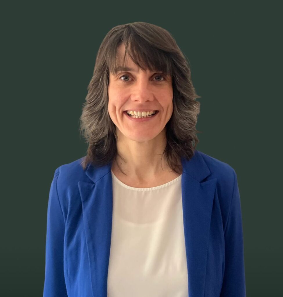 Lächelnde Frau mit dunkelbraunem, schulterlangem Haar in blauem Blazer und weißer Bluse vor dunkelgrünem Hintergrund.